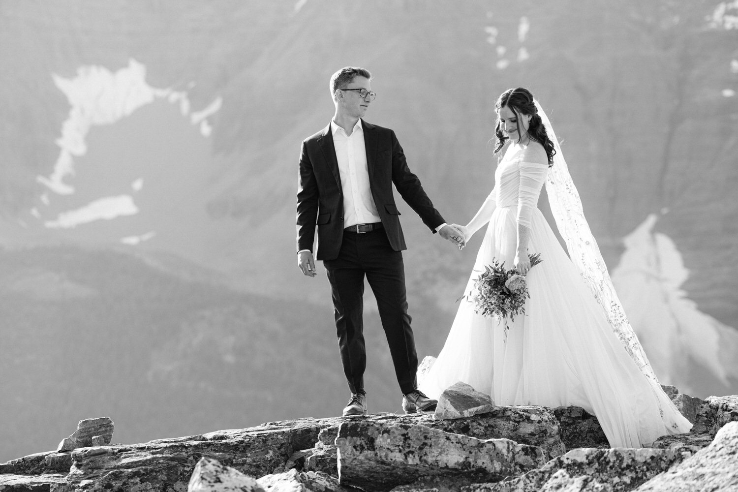 A couple holding hands on a rocky outcrop, against a stunning mountain backdrop, showcasing love in a breathtaking setting.