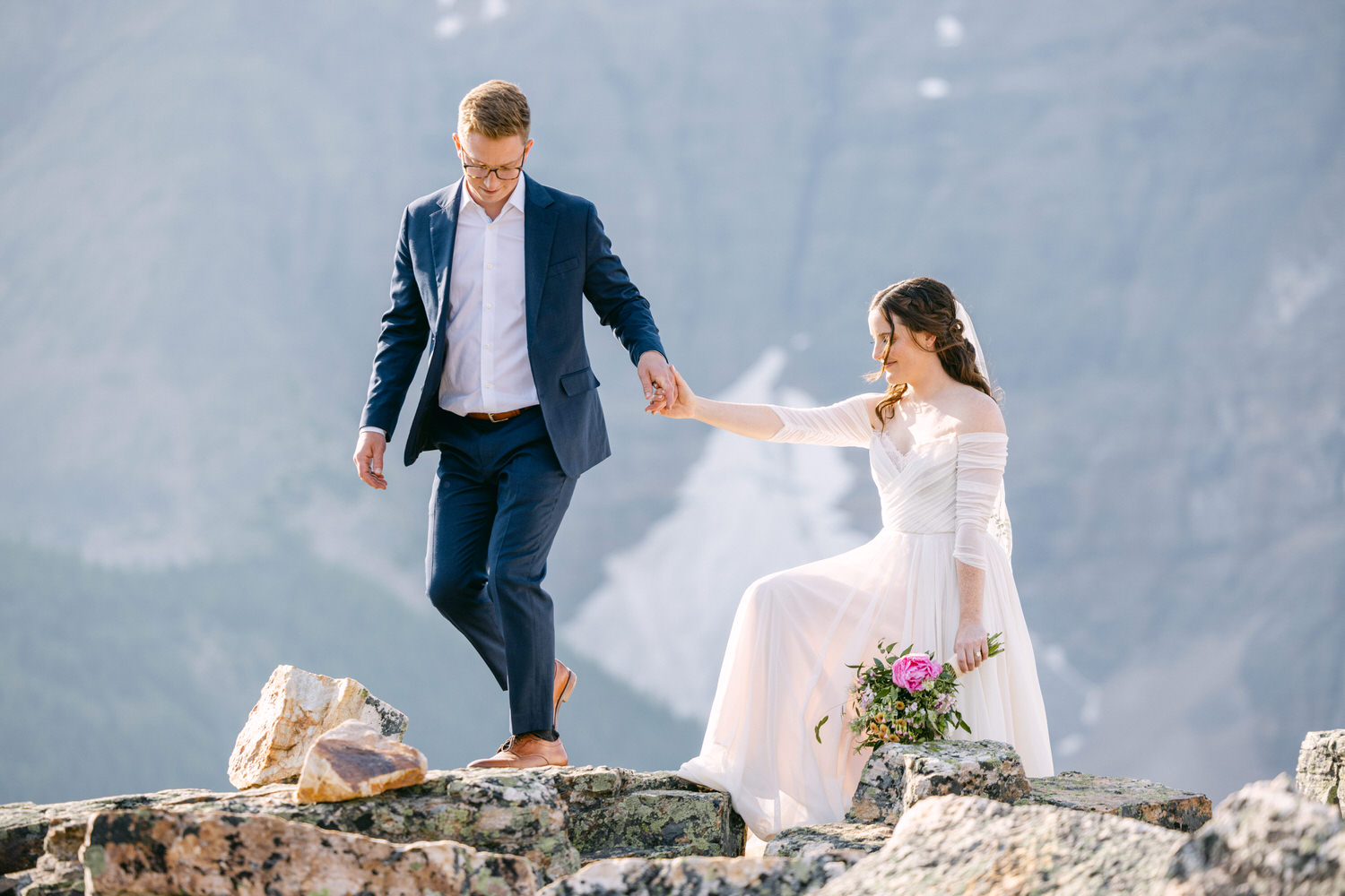 A bride and groom walking hand in hand on rocky terrain with a stunning mountain backdrop.