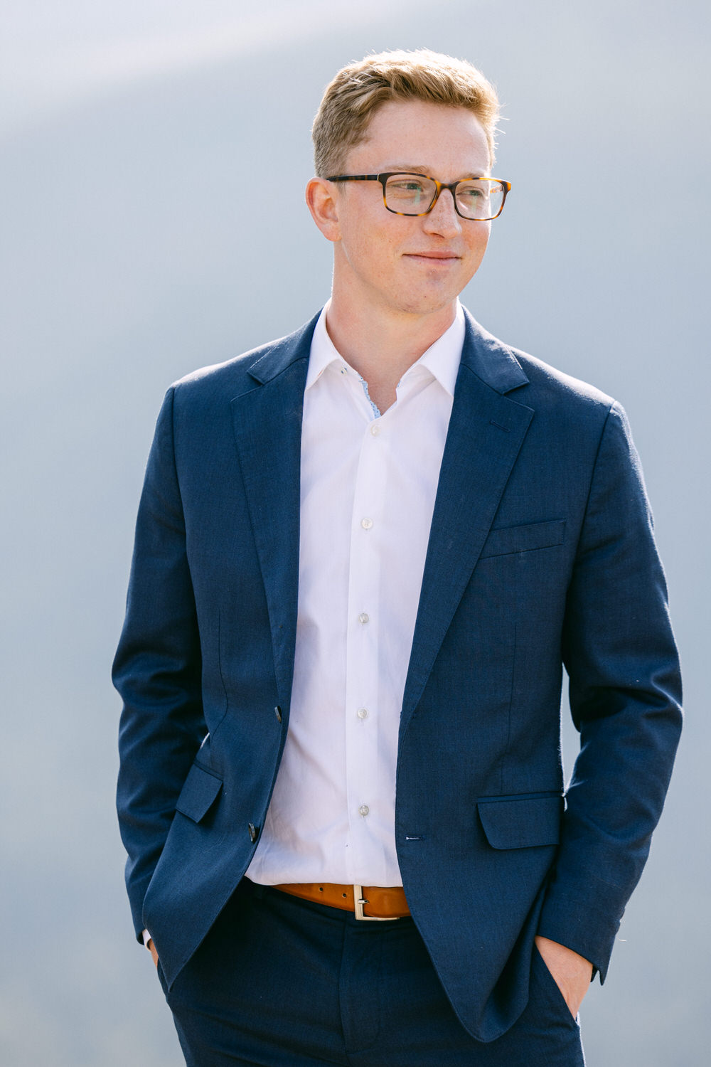 A young man wearing a navy suit and glasses stands confidently with a slight smile, hands in pockets, against a blurred outdoor background.