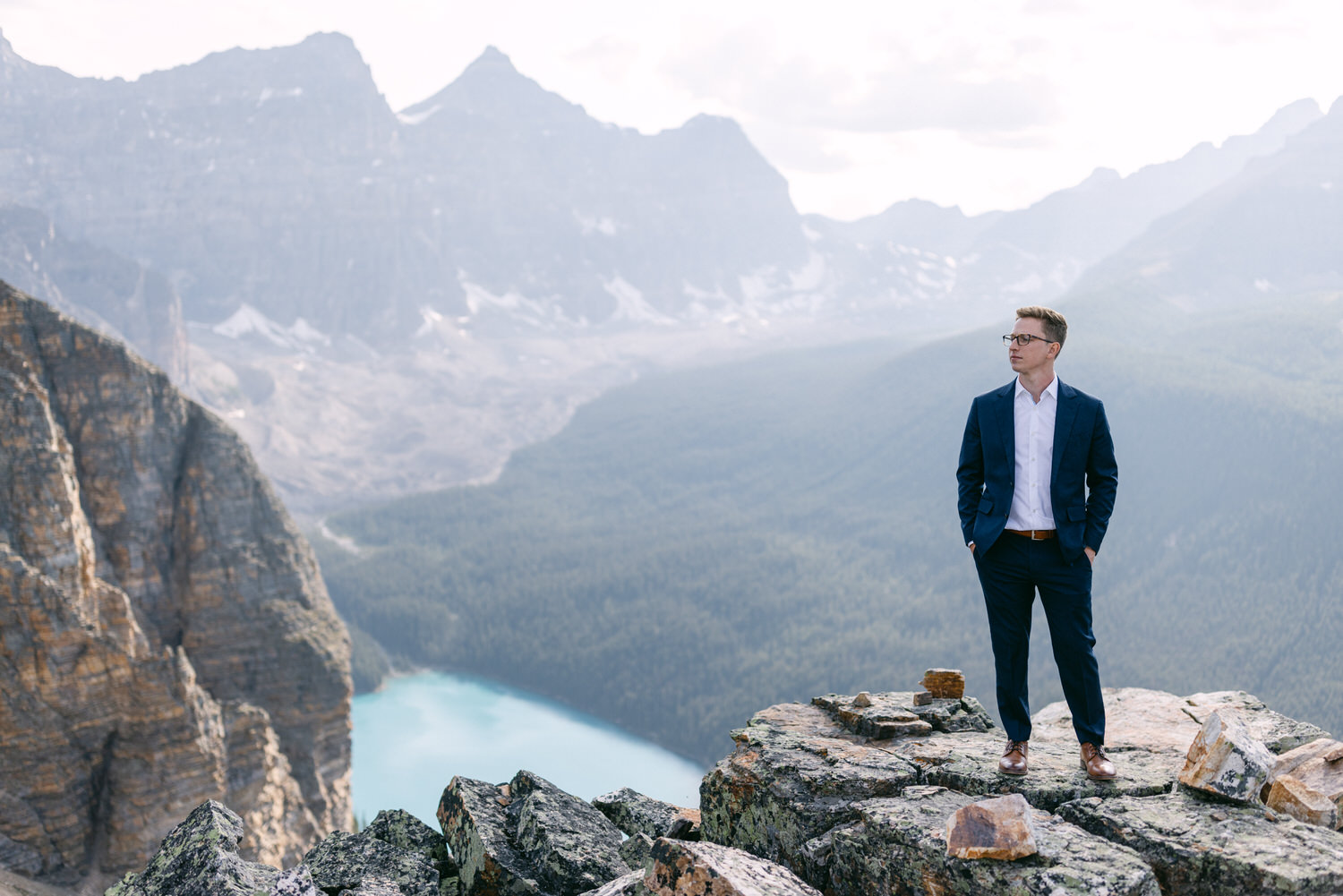 A man in a suit stands confidently on a rocky ledge overlooking a serene landscape of mountains and a blue lake.