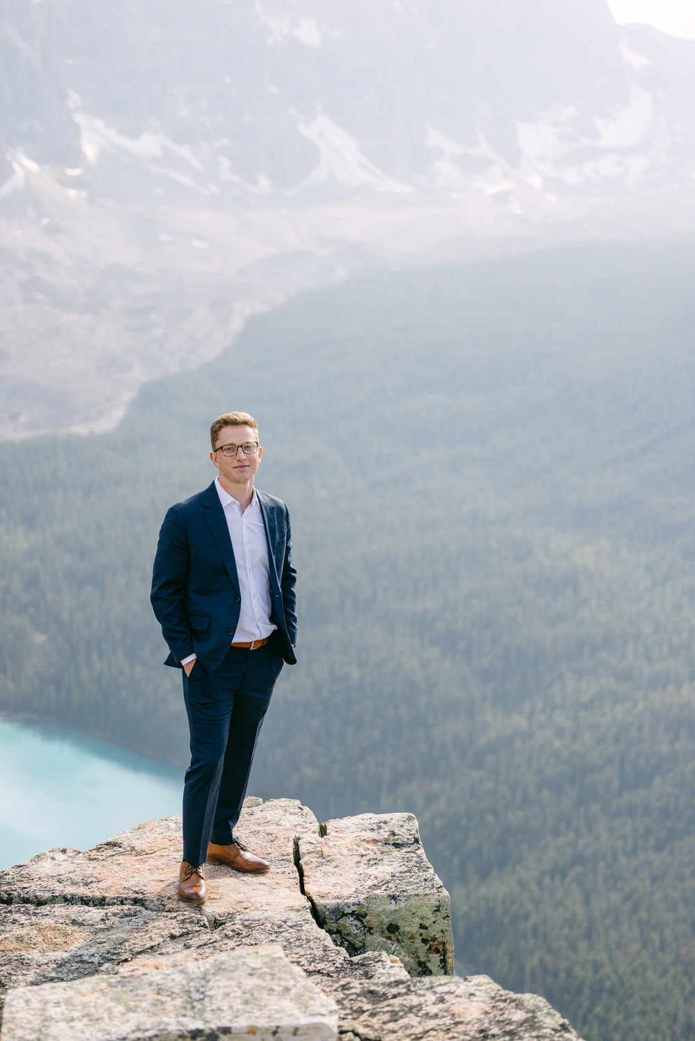 A well-dressed man stands confidently on a rocky ledge overlooking a vast forested valley and distant mountains.