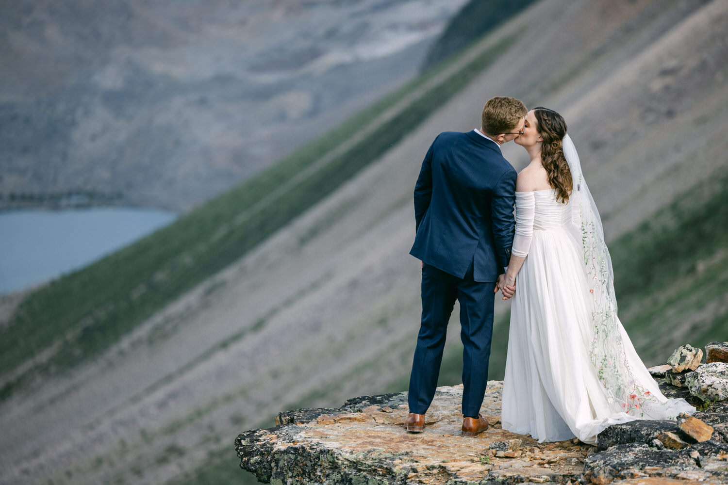 A bride and groom share a kiss while standing on a rocky outcrop overlooking a scenic landscape with mountains and a lake in the background.