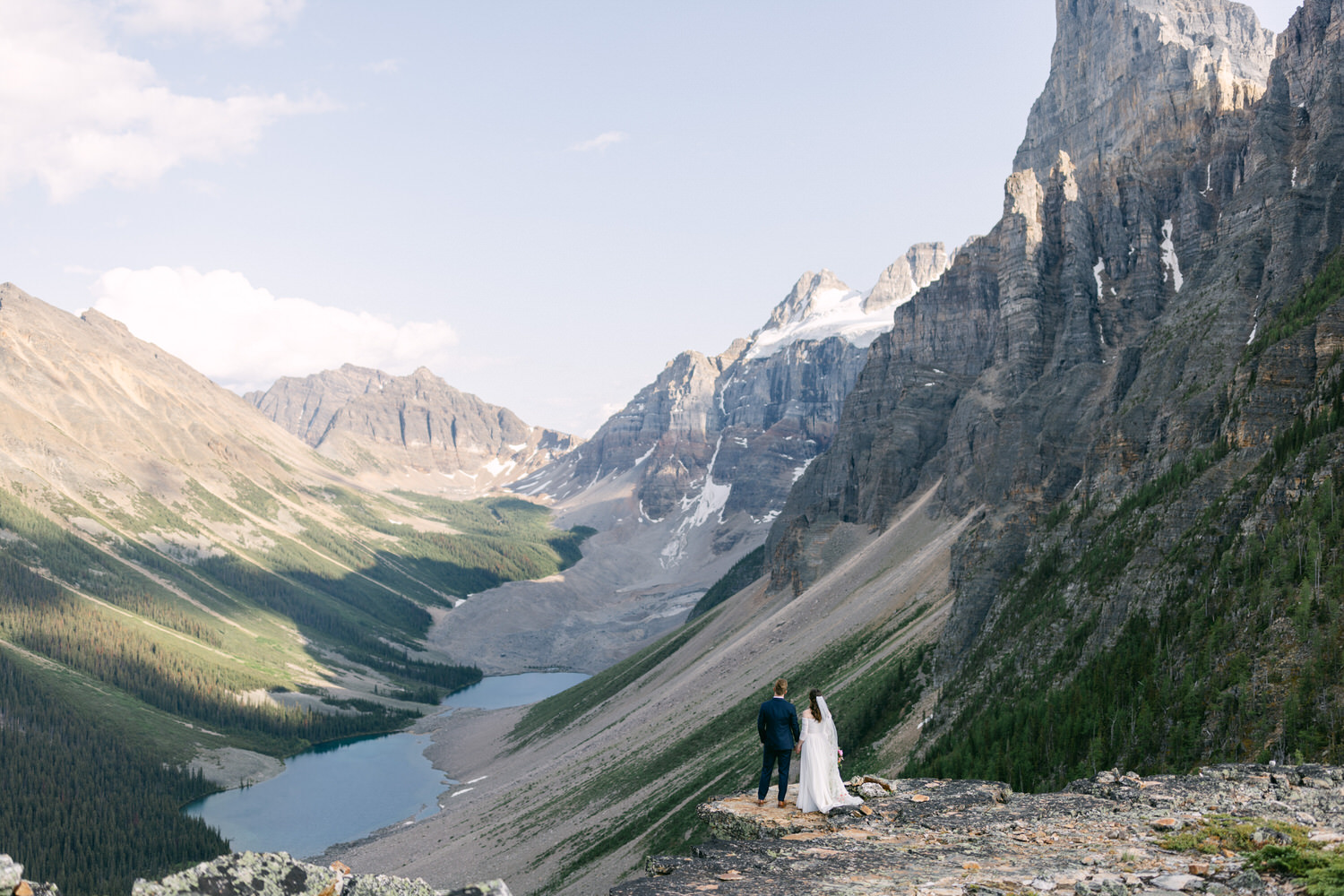 A couple stands hand-in-hand, gazing at a breathtaking mountain landscape with a serene lake below.