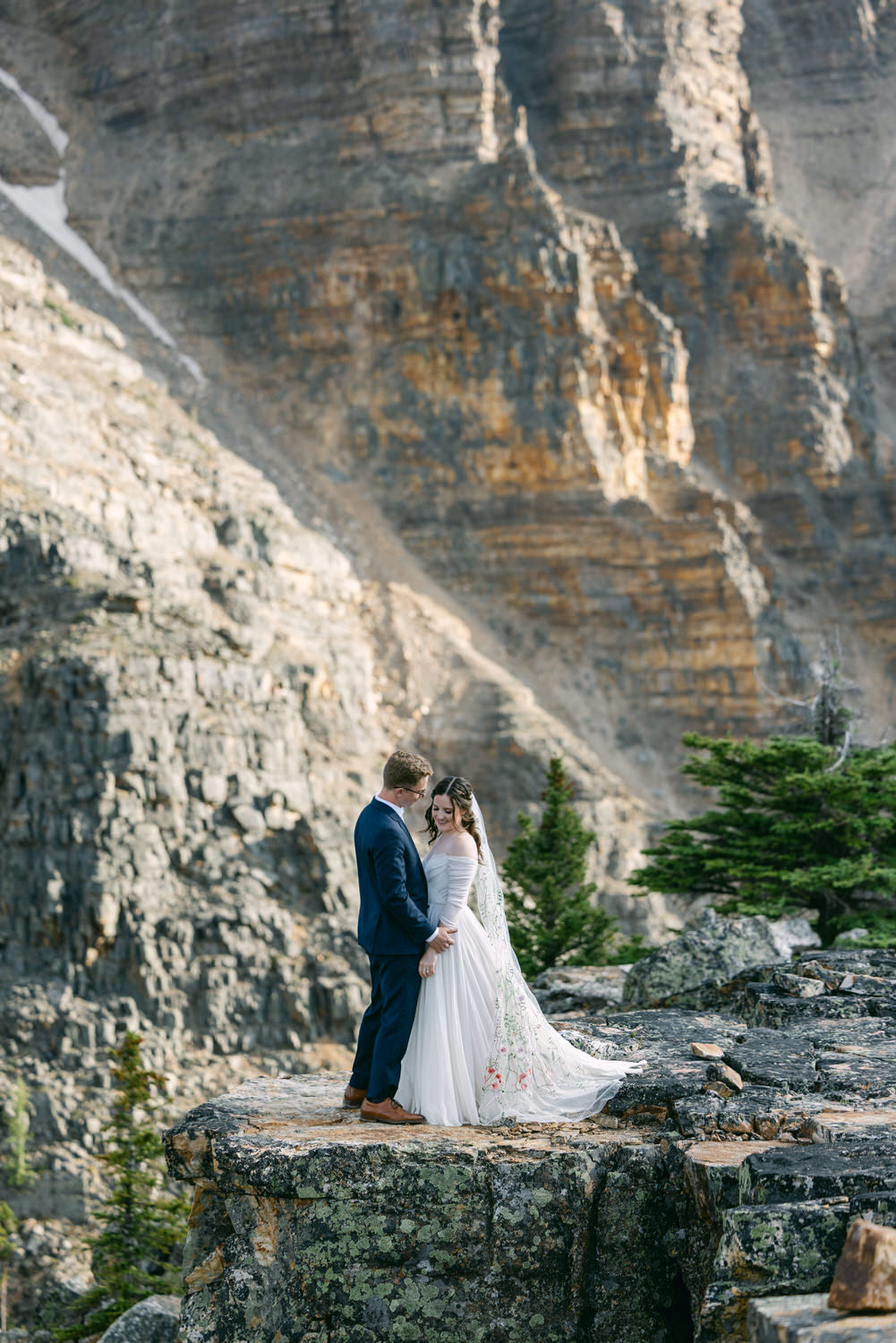 A bride and groom share a tender moment on a rocky ledge surrounded by towering mountains and greenery.