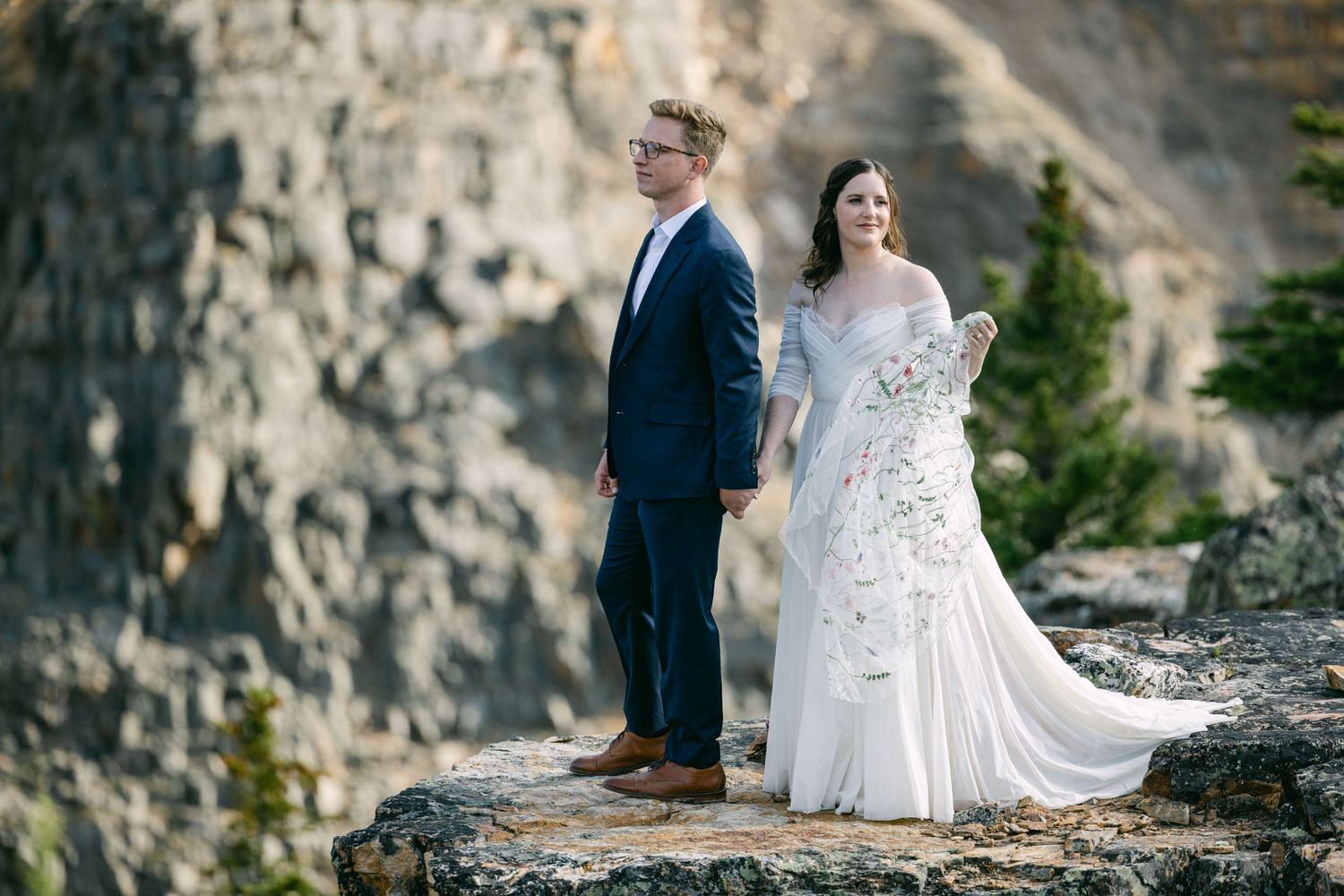 Bride and groom holding hands on a rocky ledge with mountains in the background.