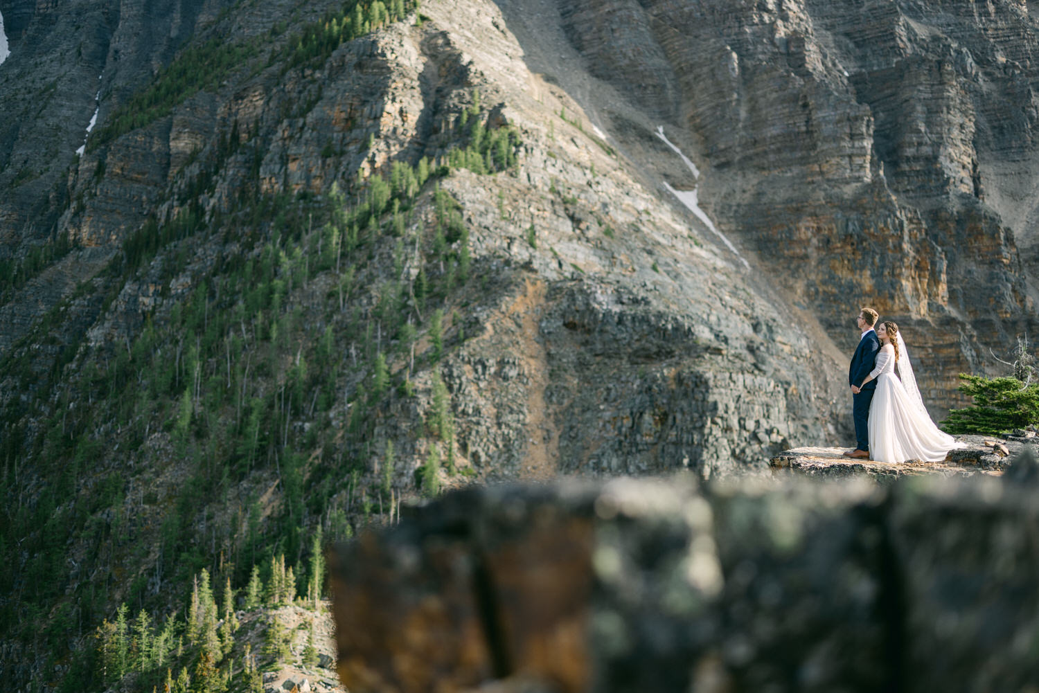 A couple stands hand in hand on a rocky ledge, surrounded by majestic mountains and greenery.