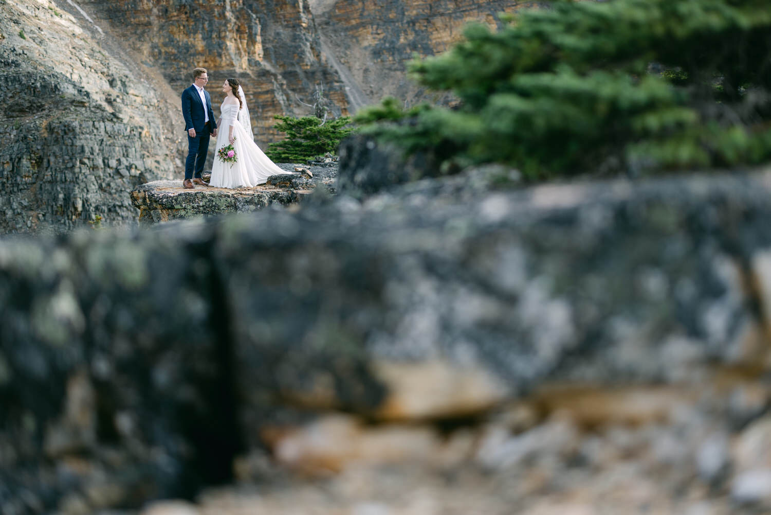 A bride and groom stand on a rocky ledge surrounded by natural beauty, capturing a moment of love amidst the landscape.