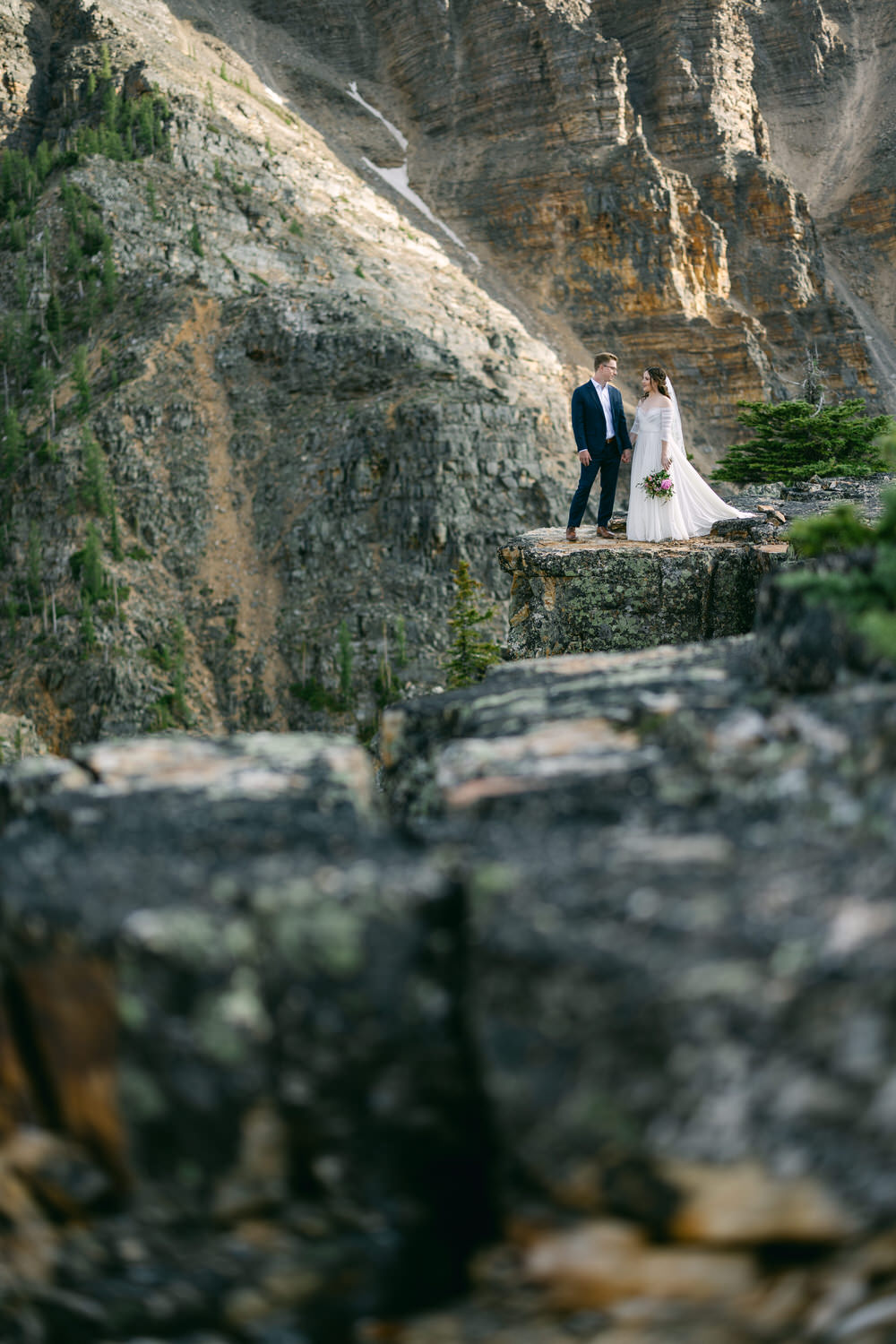 A couple stands together on a rocky ledge, surrounded by majestic mountains, celebrating their love in a breathtaking outdoor setting.