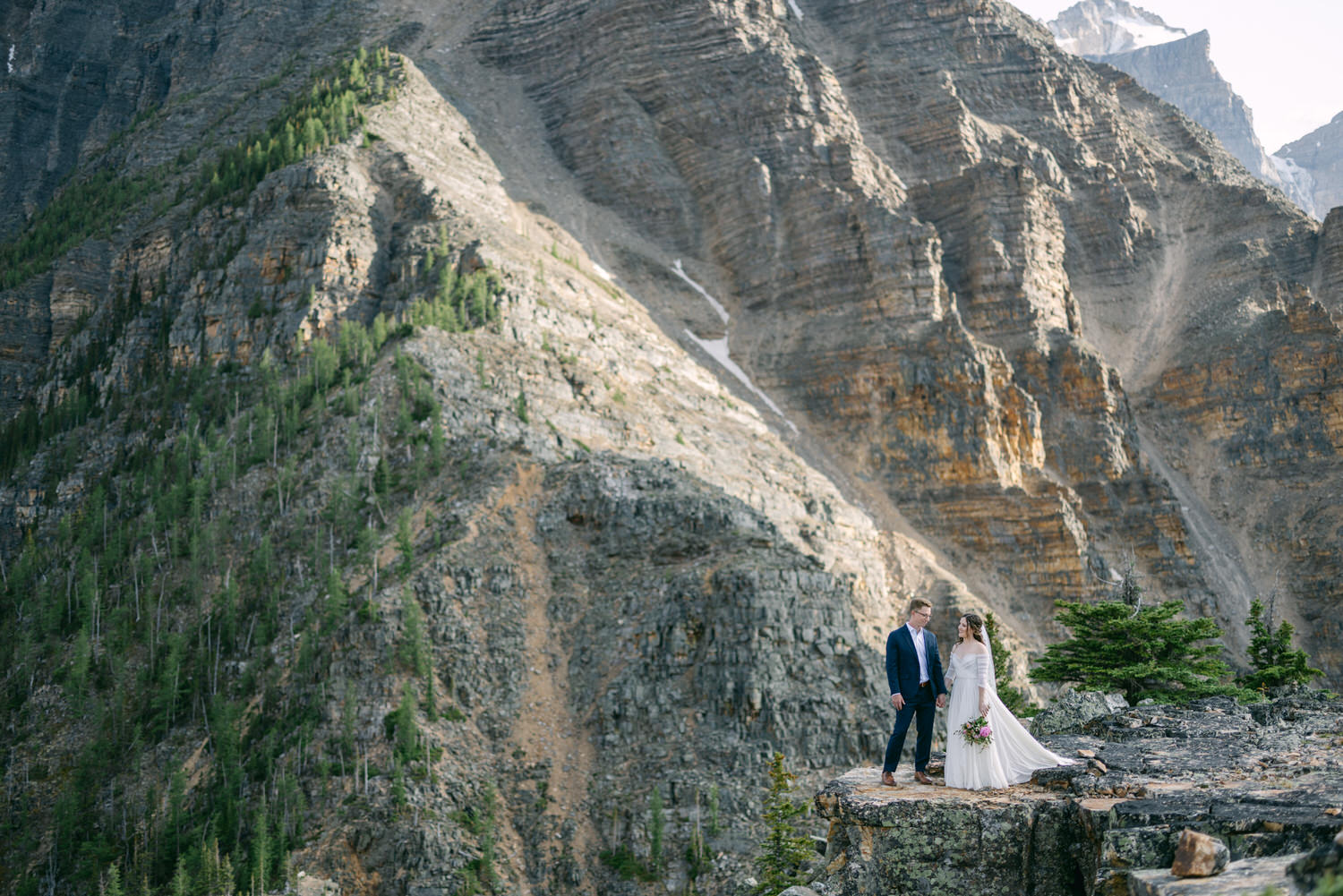 A couple stands on a rocky outcrop surrounded by majestic mountains, celebrating their wedding in a breathtaking outdoor setting.