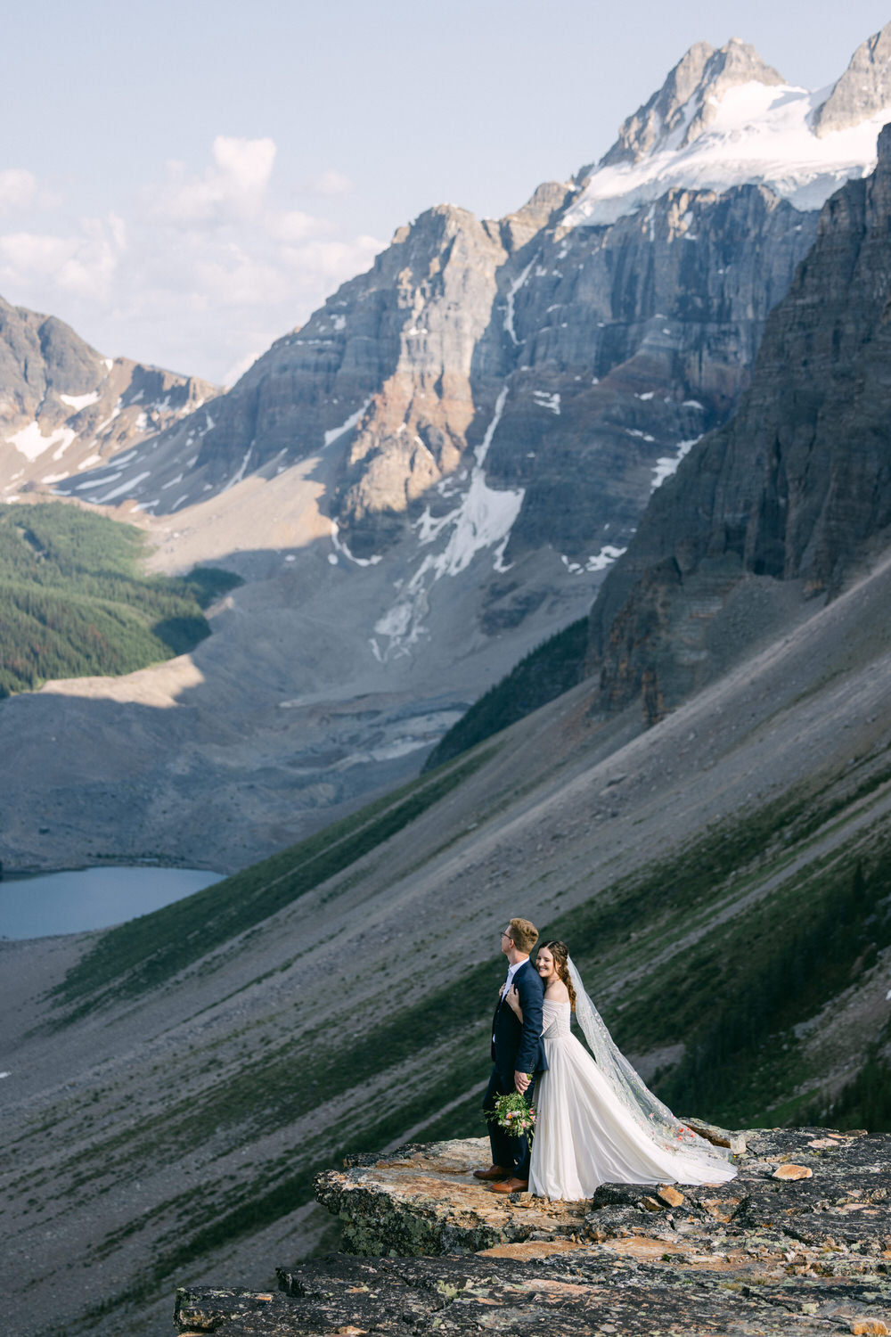 A couple stands on a rocky outcrop, surrounded by majestic mountains and a serene lake, celebrating their wedding day in a stunning natural setting.