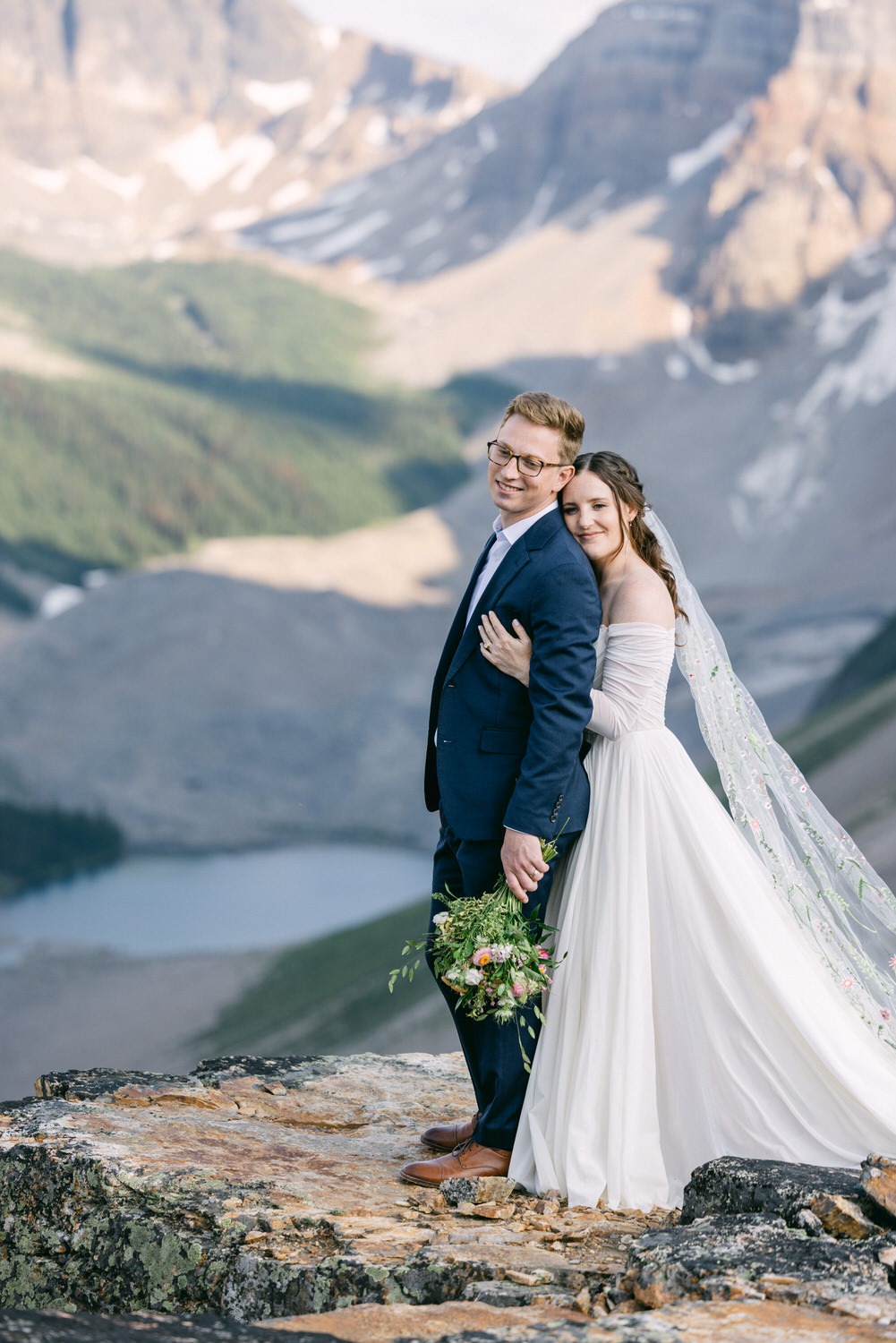 A bride and groom embrace on a rocky outcrop with a mountainous landscape in the background, showcasing their wedding attire and surroundings.