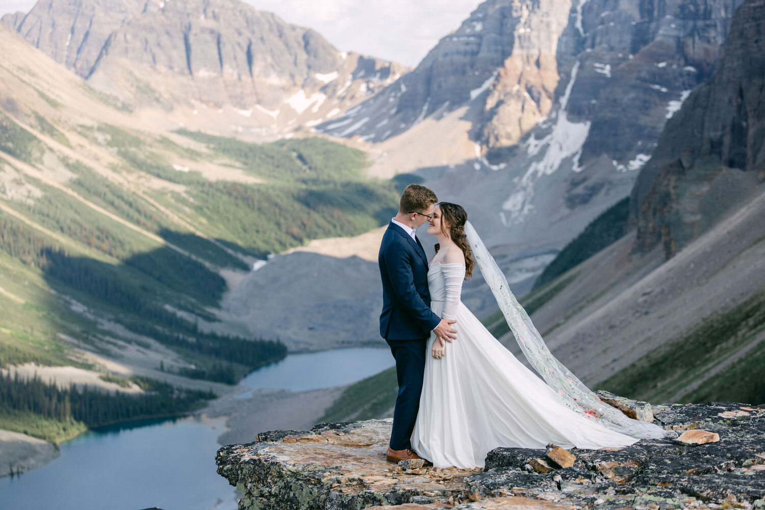 A happy couple shares a romantic kiss in formal attire, with a breathtaking mountain landscape and serene lake in the background.