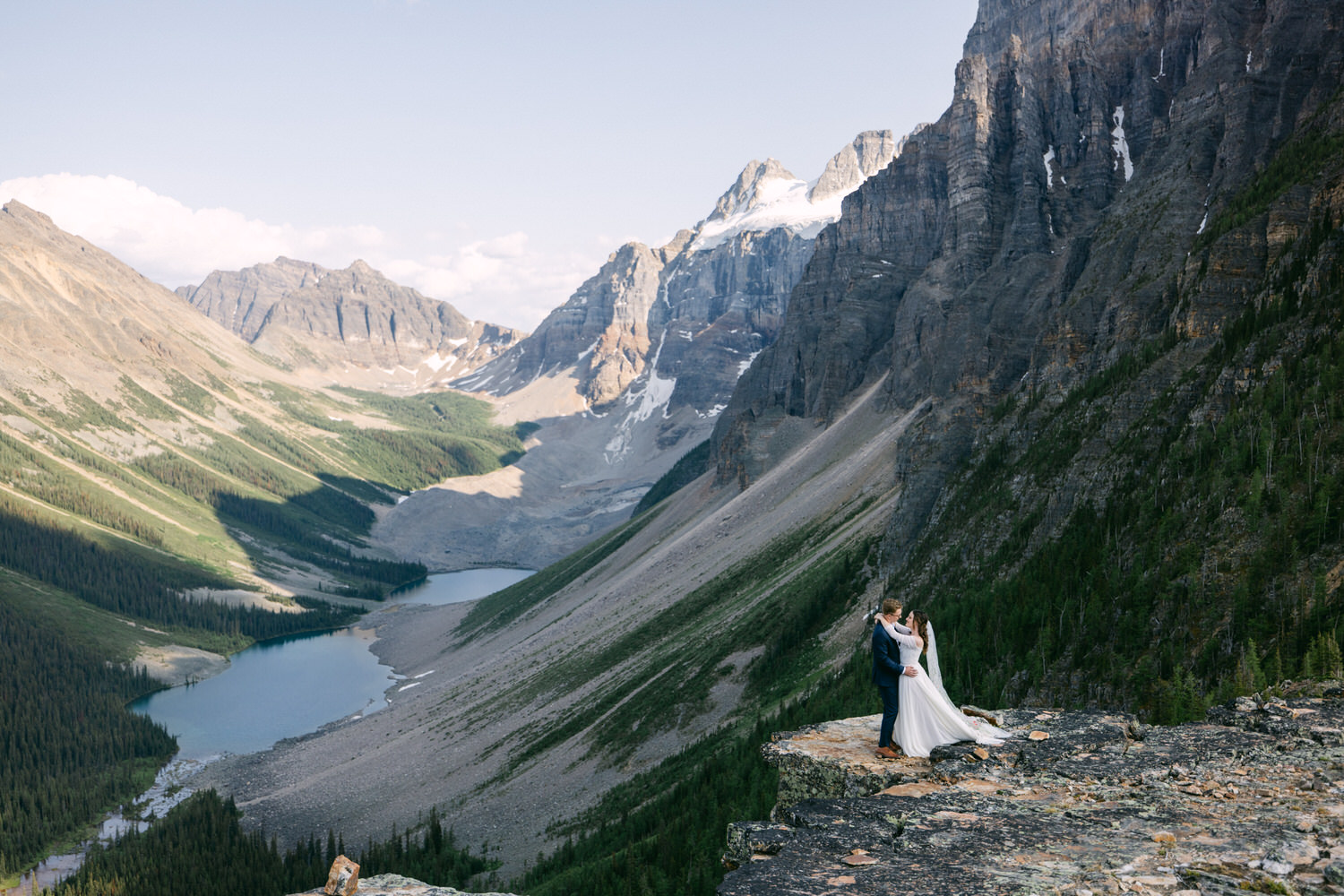 A couple sharing an intimate moment on a rocky outcrop overlooking a scenic valley and lake, surrounded by majestic mountains.