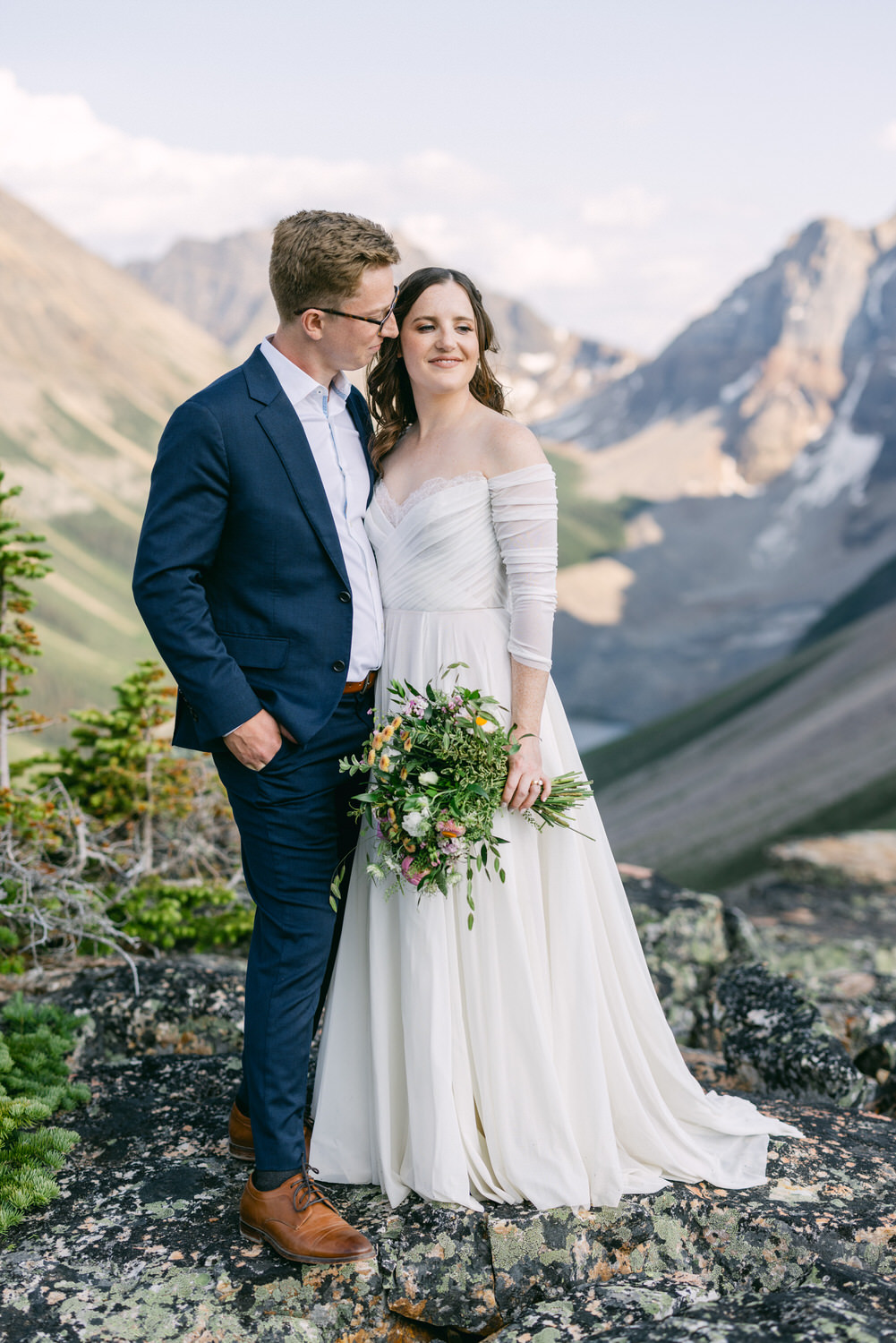 A couple shares an intimate moment in a stunning mountain landscape, the bride holding a bouquet of flowers, dressed in an elegant white gown, while the groom is in a smart suit.