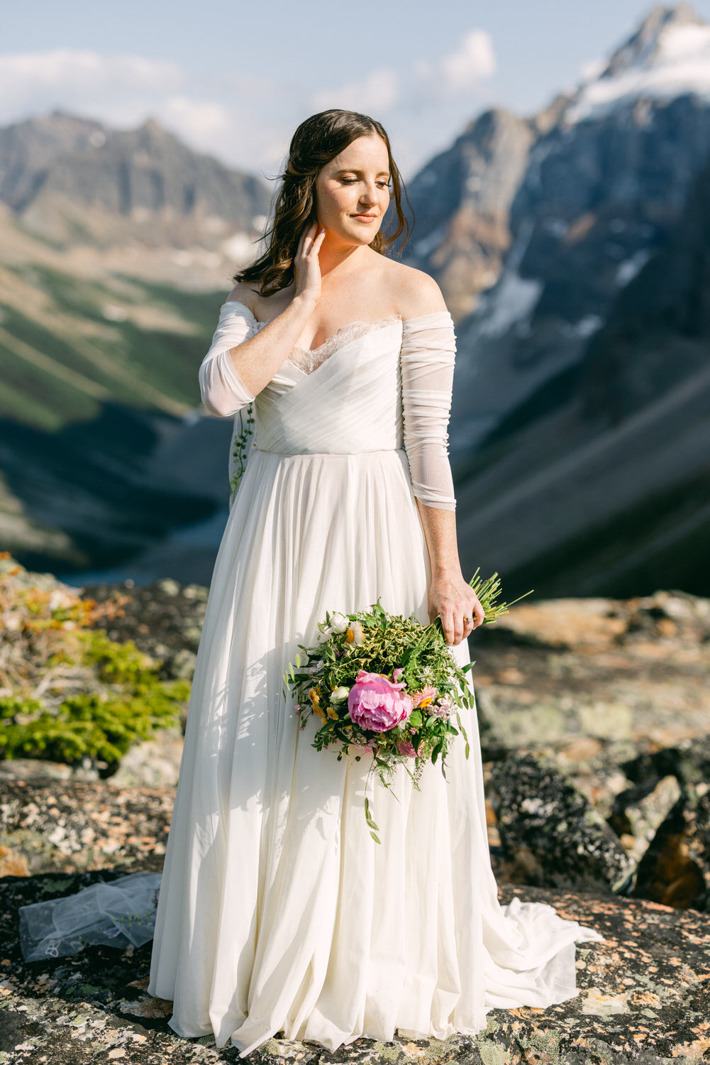 A bride in a flowing white gown poses gracefully outdoors, holding a vibrant floral bouquet against a stunning mountainous backdrop.