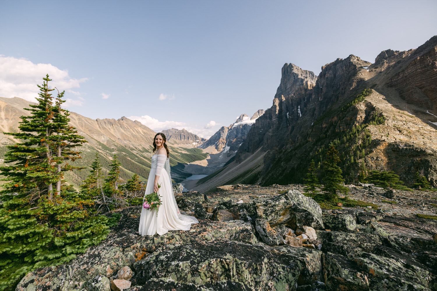 A bride in a flowing gown stands on a rocky outcrop with majestic mountains in the background, holding a bouquet of flowers amidst a scenic landscape.