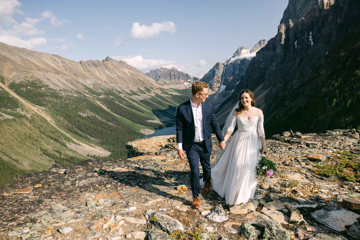 A couple walks hand-in-hand on a rocky mountain ledge, surrounded by stunning landscapes, as the bride holds a bouquet.