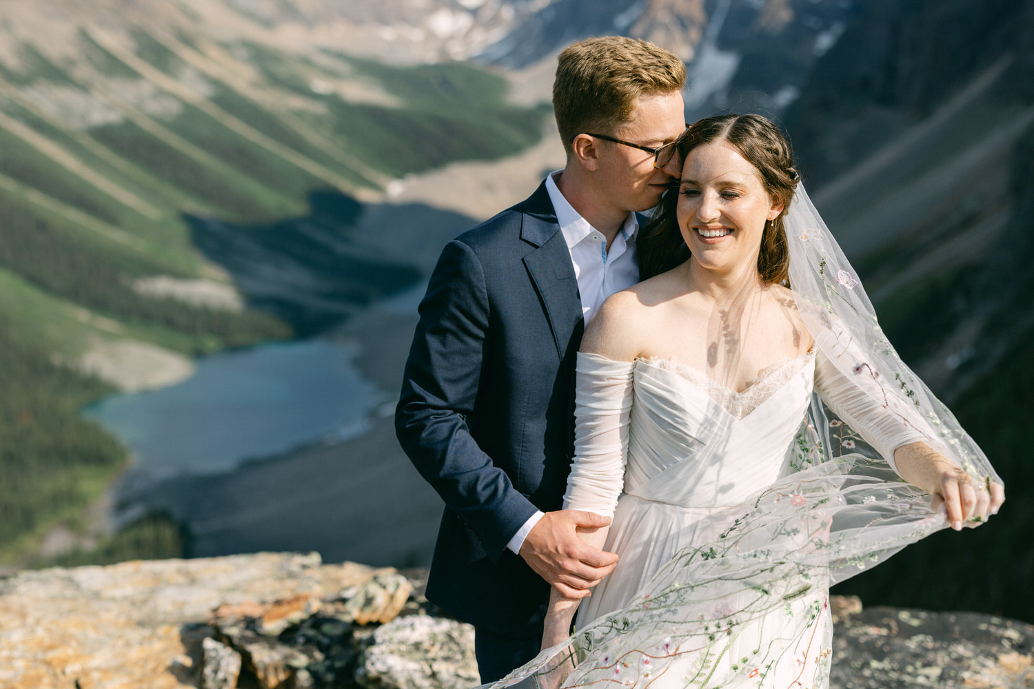 A smiling couple shares a tender moment on a dramatic mountain overlook, with a stunning landscape of green valleys and a shimmering lake in the background.