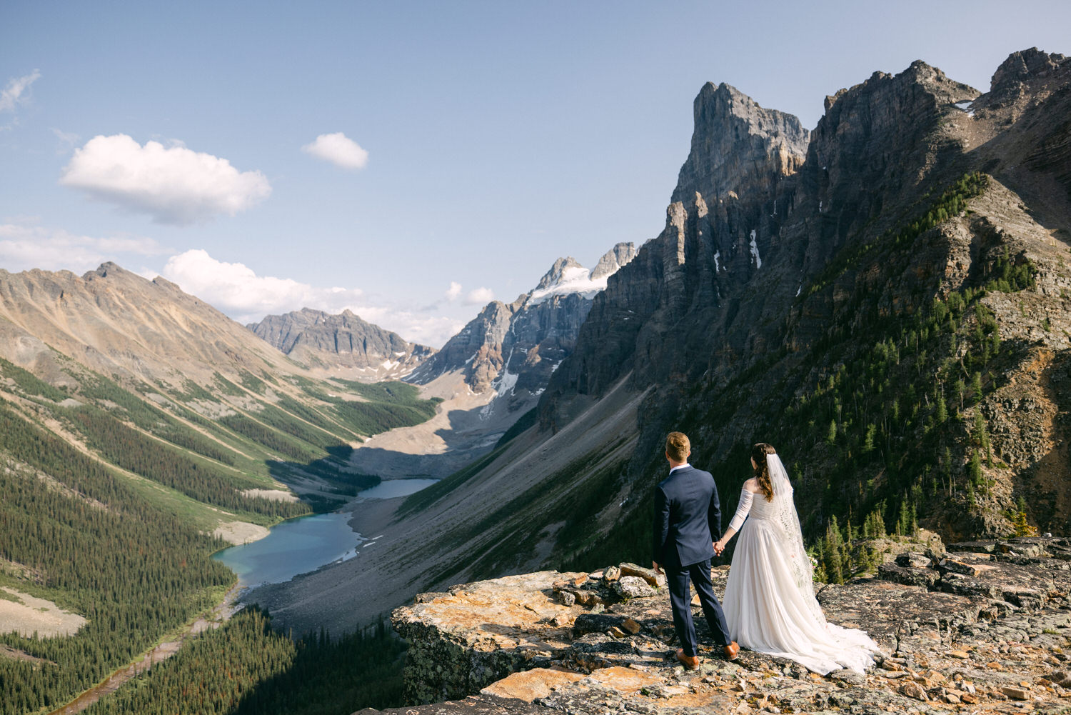 A couple stands hand in hand, overlooking a breathtaking mountain landscape, with lush greenery and a tranquil lake below.