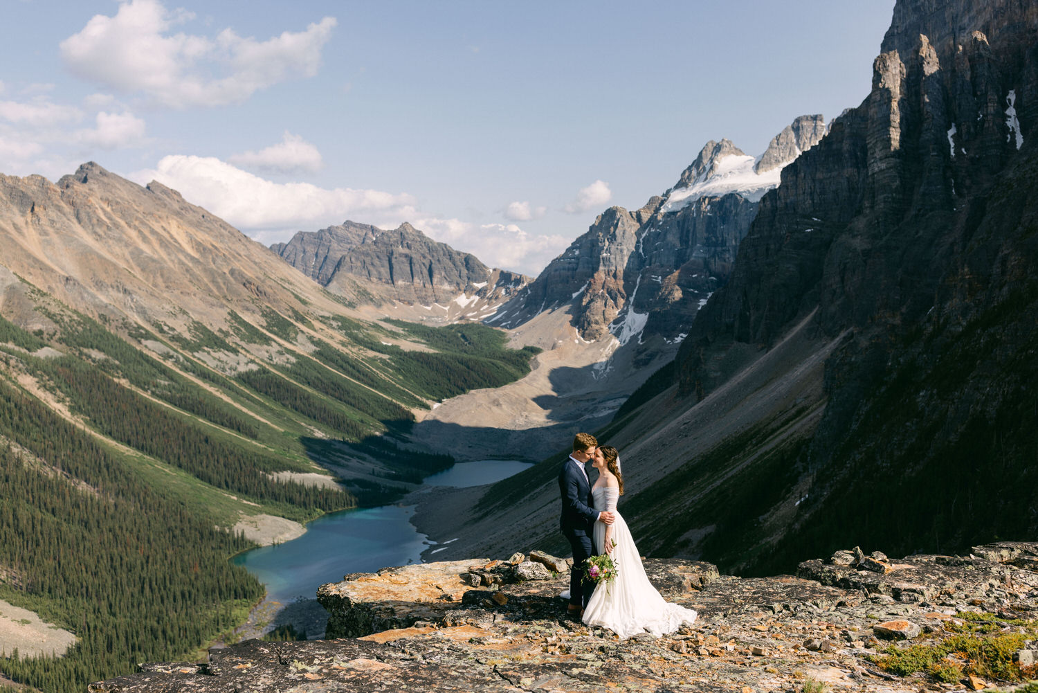 A bride and groom share a kiss against a stunning mountain backdrop, surrounded by a lush valley and a clear blue lake.