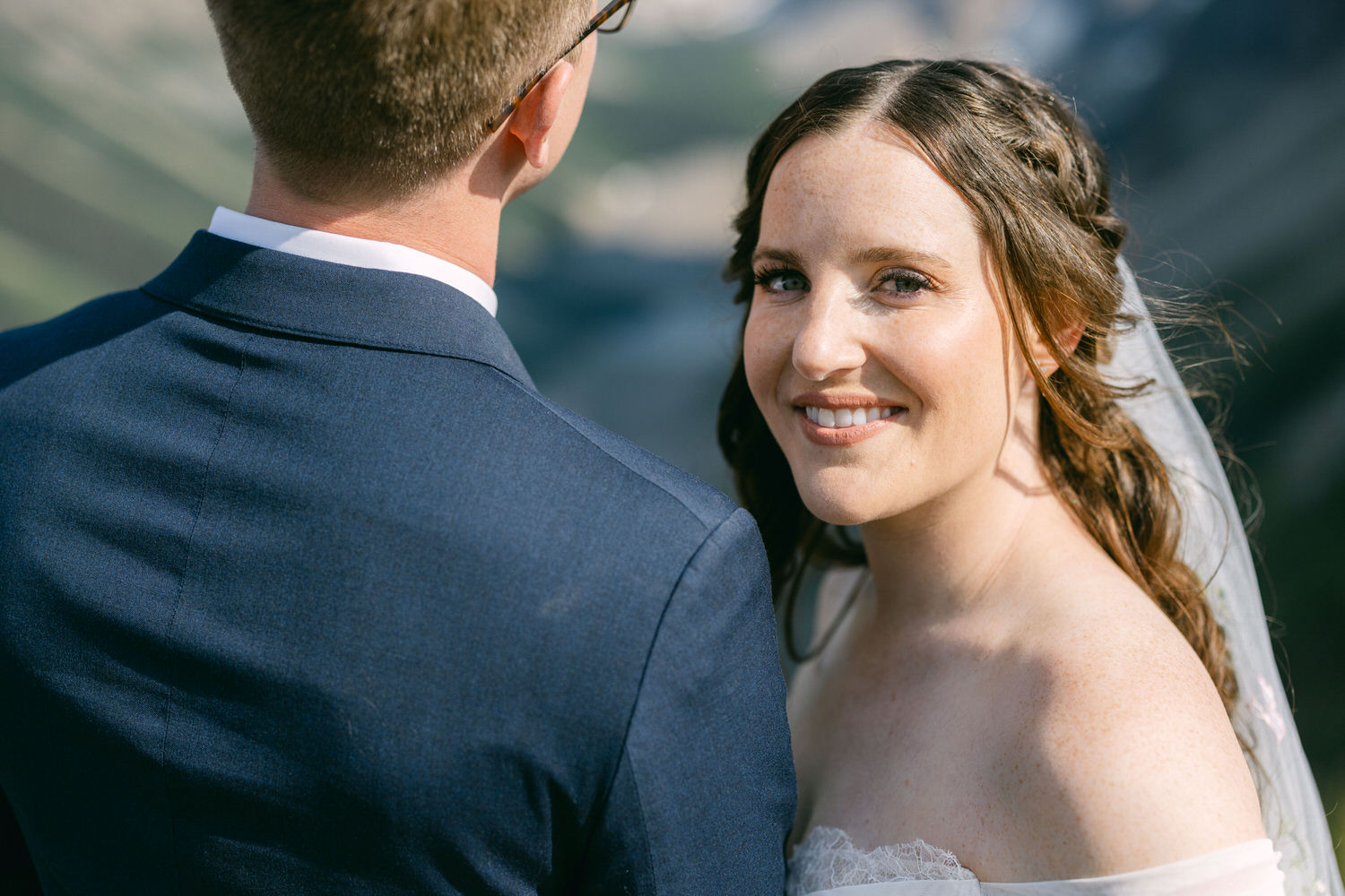 A bride smiles warmly at the camera with her groom's back facing her, amidst a beautiful outdoor setting.