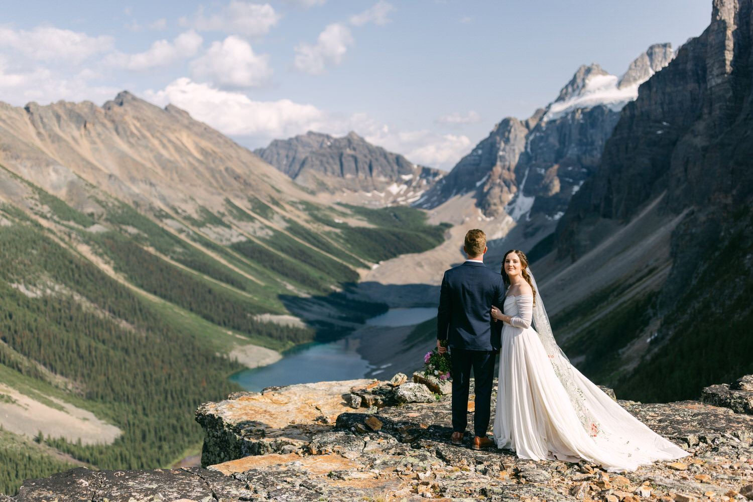 A couple stands on a rocky ledge overlooking a breathtaking mountain landscape, radiating love in a serene outdoor wedding setting.