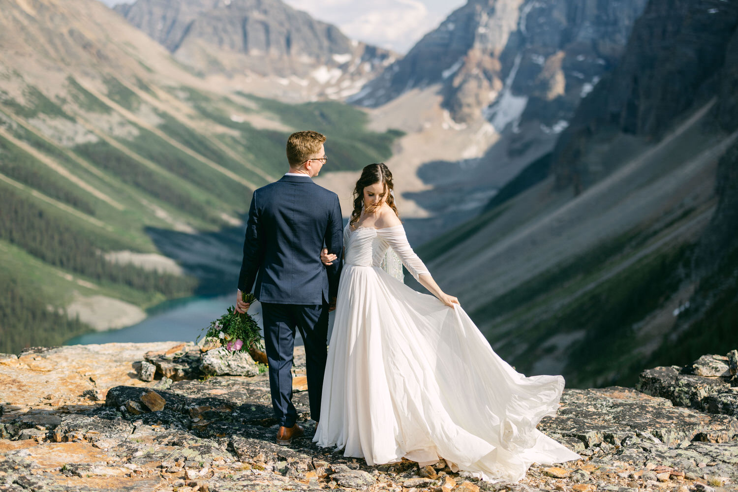 A couple stands hand in hand overlooking a stunning mountain landscape, with the bride in a flowing white dress and the groom in a navy suit.