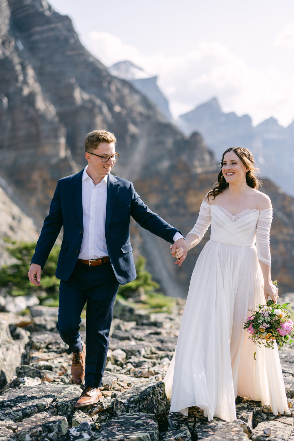 A newlywed couple holding hands while walking on rocky terrain, with a breathtaking mountain backdrop, showcasing joy and romance.