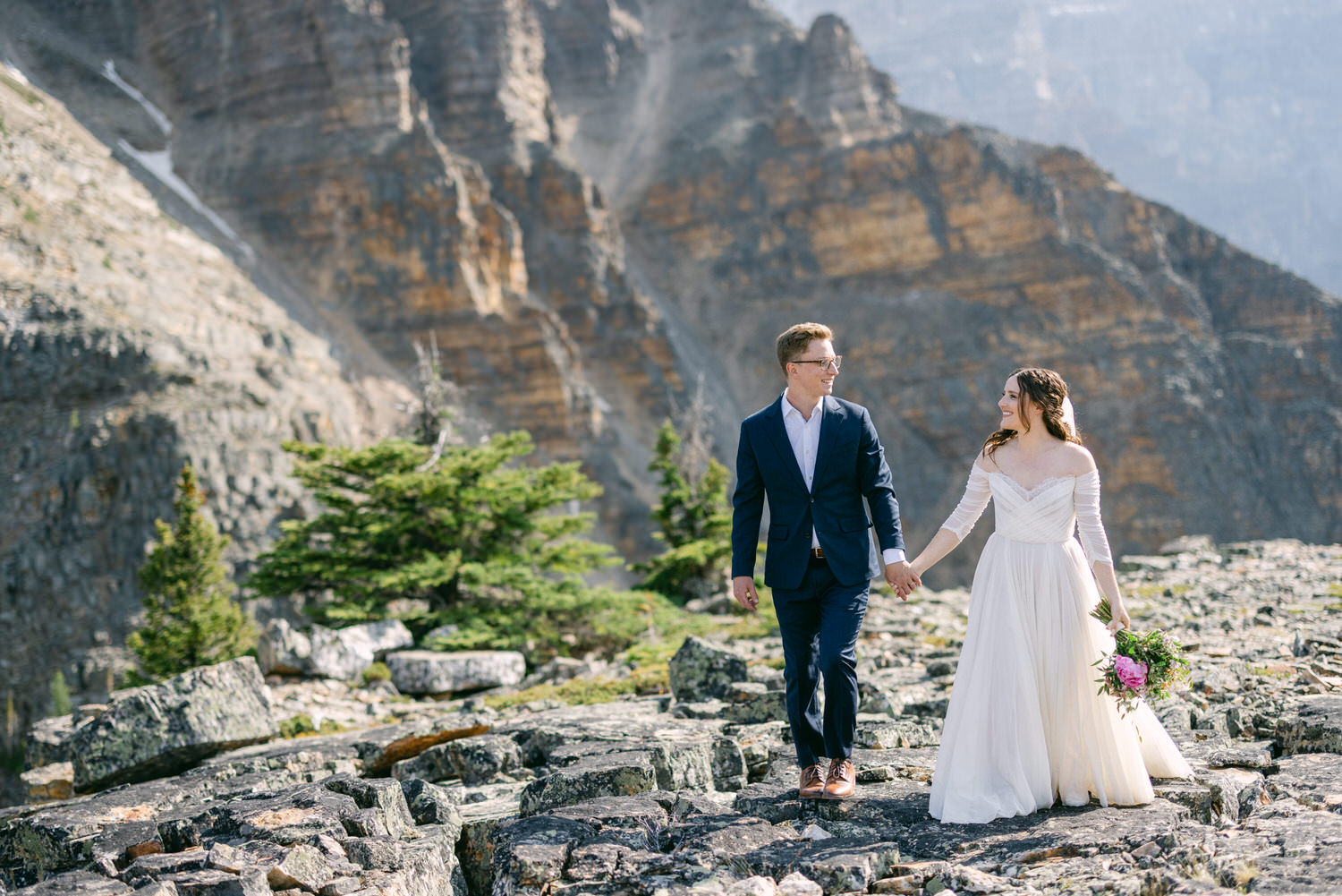 A bride in a flowing white gown and a groom in a dark suit hold hands while walking on rocky terrain, surrounded by mountains and evergreen trees.