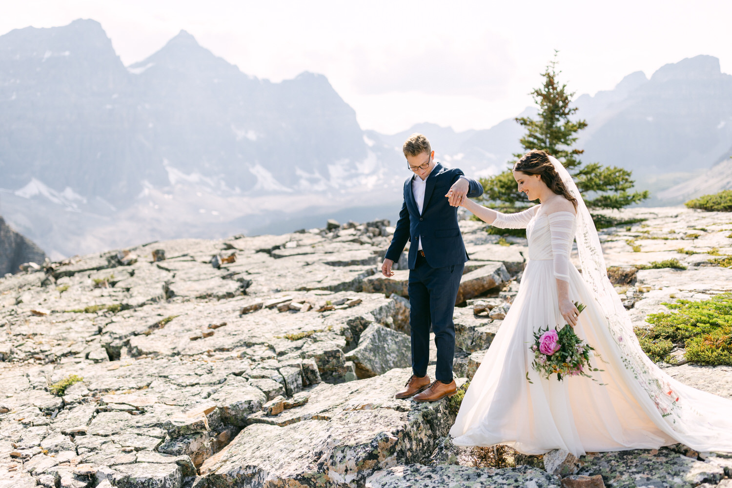 A couple shares a romantic moment as they navigate rocky terrain during their wedding amidst stunning mountain scenery.