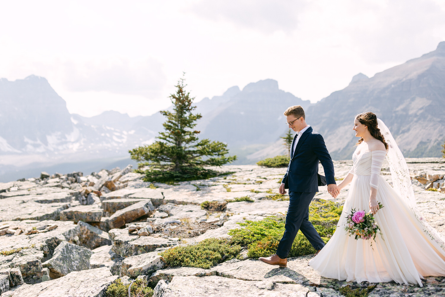 A bride and groom walk hand-in-hand across rocky terrain with mountains in the background, showcasing a scenic outdoor wedding moment.