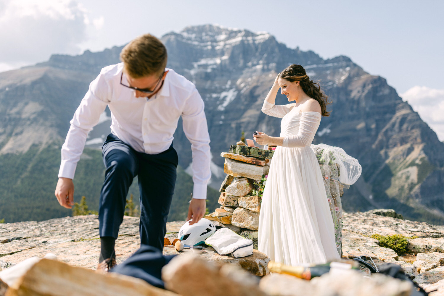 A couple prepares for a mountain wedding, with the bride in a flowing white dress and the groom adjusting his attire, surrounded by stunning alpine scenery.