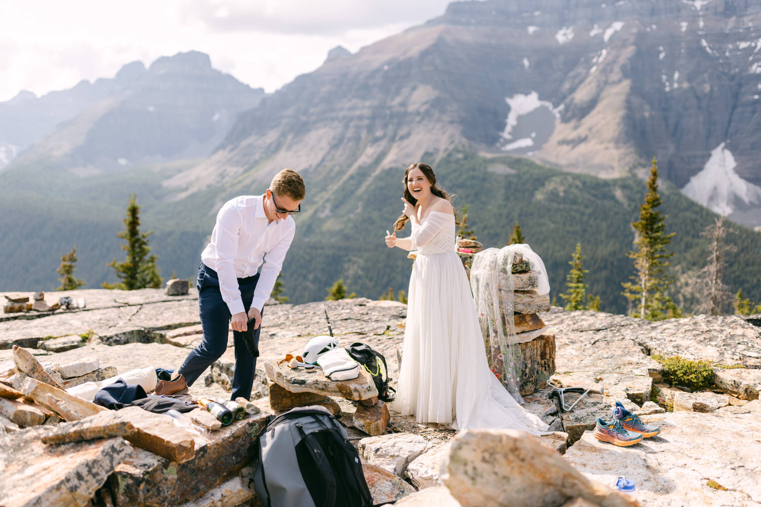 A couple preparing for their wedding in a scenic mountain setting, with the bride in a flowing white dress and the groom adjusting his clothes amidst their gear and stunning nature backdrop.