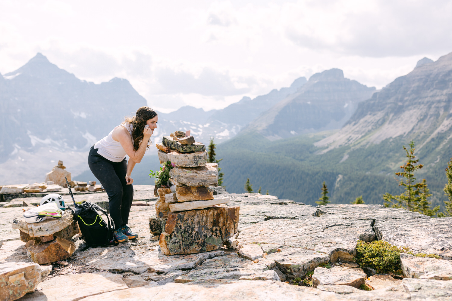 A woman in athletic wear leans over a stacked rock formation with mountains in the background, engaging with nature while exploring the outdoors.