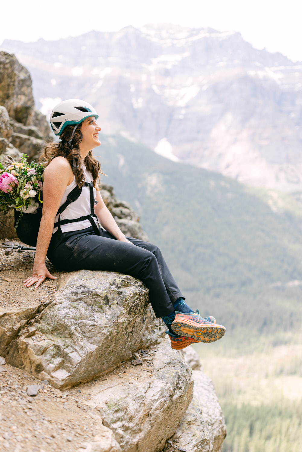 A smiling woman wearing a helmet and sitting on a rock ledge with a bouquet of flowers on her back, enjoying the scenic mountain landscape.