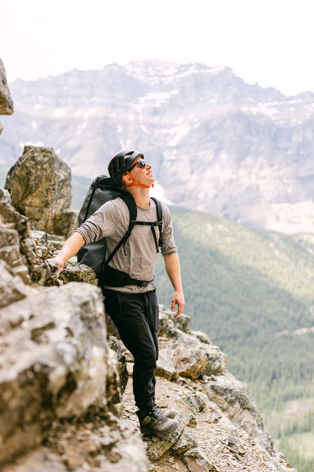 A man wearing a helmet and sunglasses stands on a rocky ledge, looking up at the mountains with a backpack, surrounded by lush green valleys.