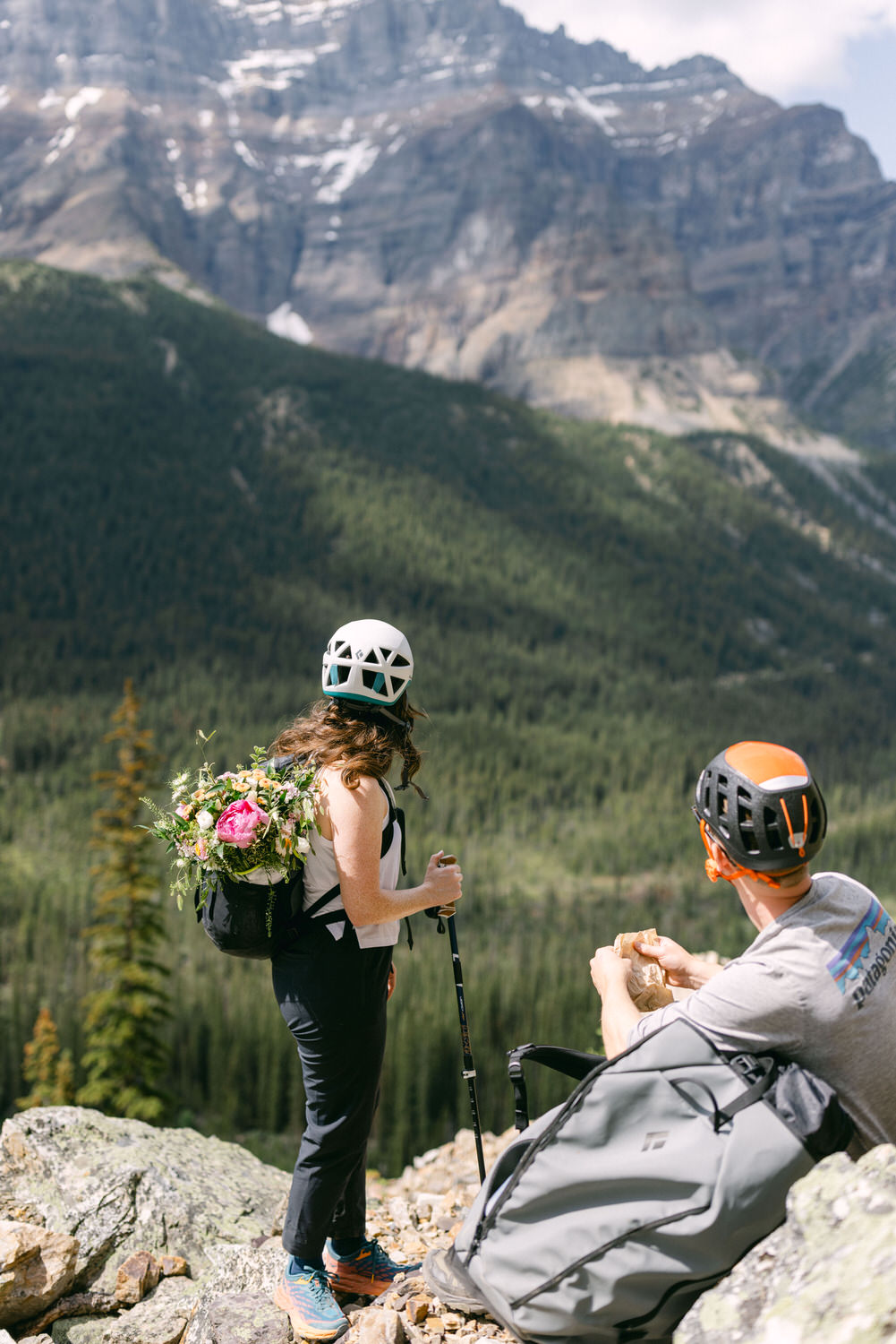 A woman with a floral backpack stands on a rocky ledge, gazing at a mountainous landscape, while a man sits nearby, eating lunch.