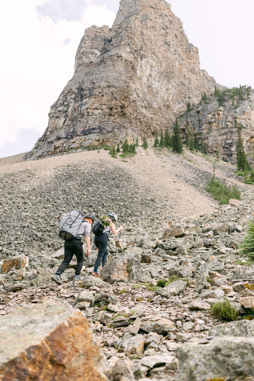 A couple navigates a steep, rocky slope on a mountain trail, surrounded by towering peaks and sparse greenery.