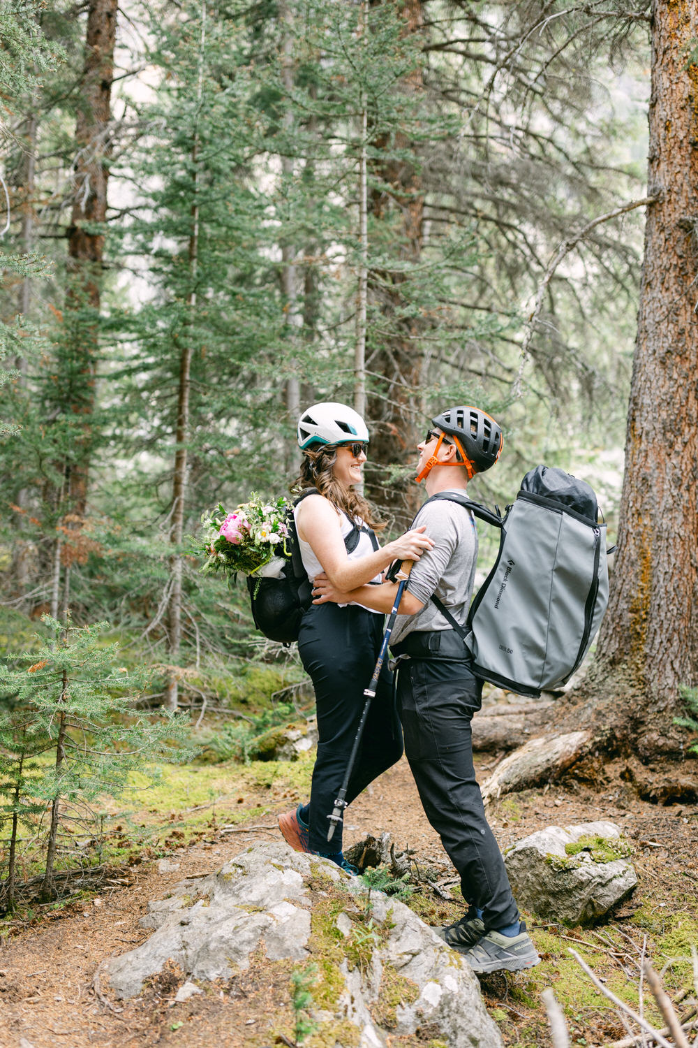 A joyful couple embraces in the woods while wearing cycling helmets and backpacks, with a bouquet of flowers in one backpack.