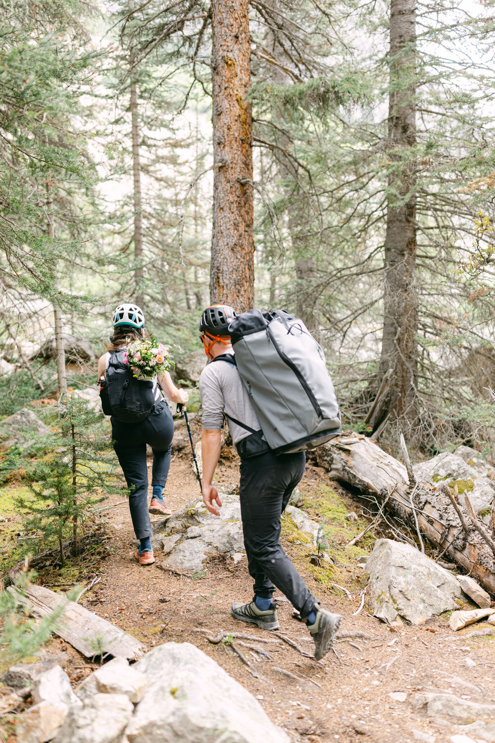 A couple hiking through a forest trail, one carrying a floral arrangement on her backpack and the other carrying a large backpack, surrounded by trees and rocks.