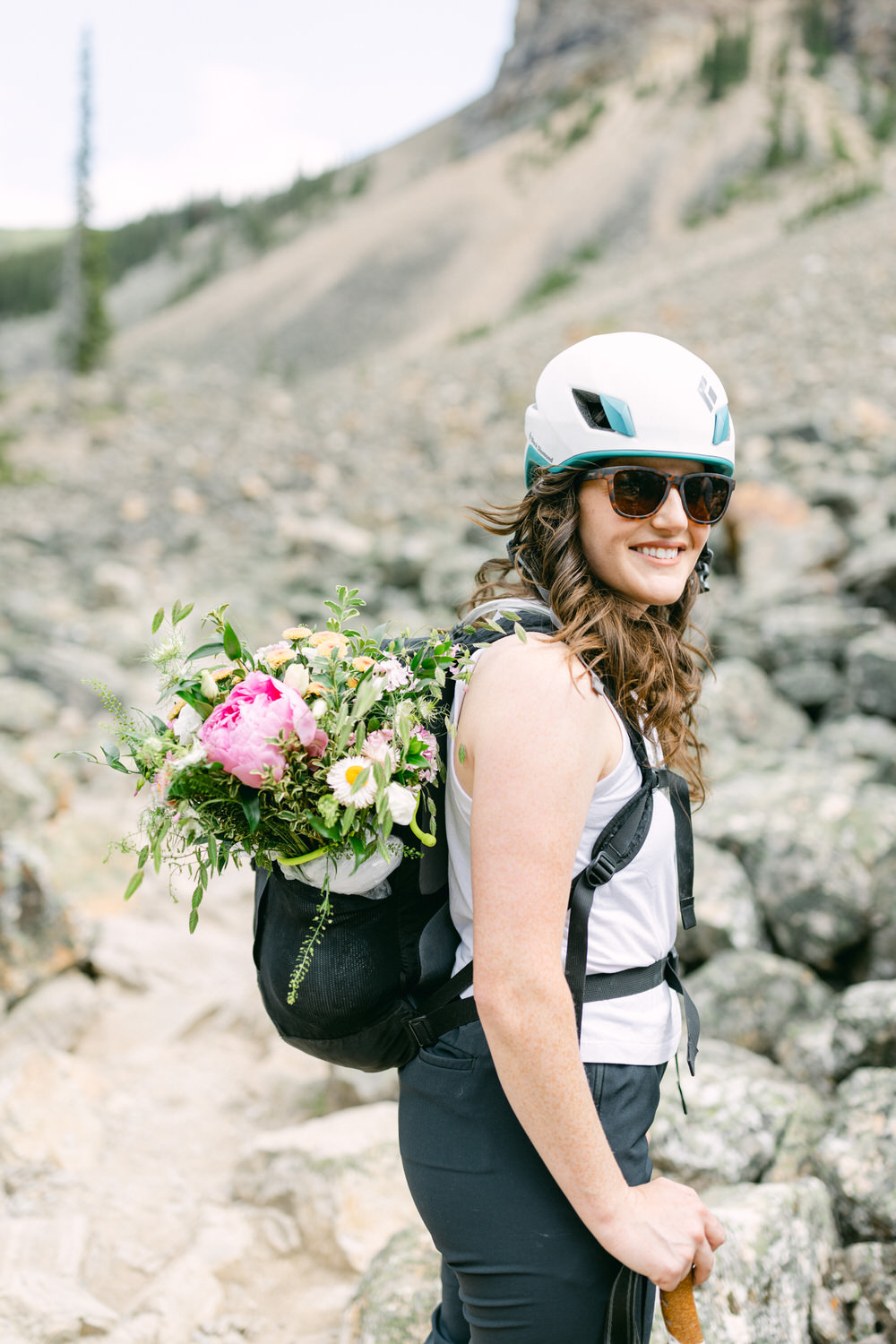 A woman wearing a helmet and sunglasses smiles while hiking on a rocky trail, carrying a backpack filled with vibrant flowers.