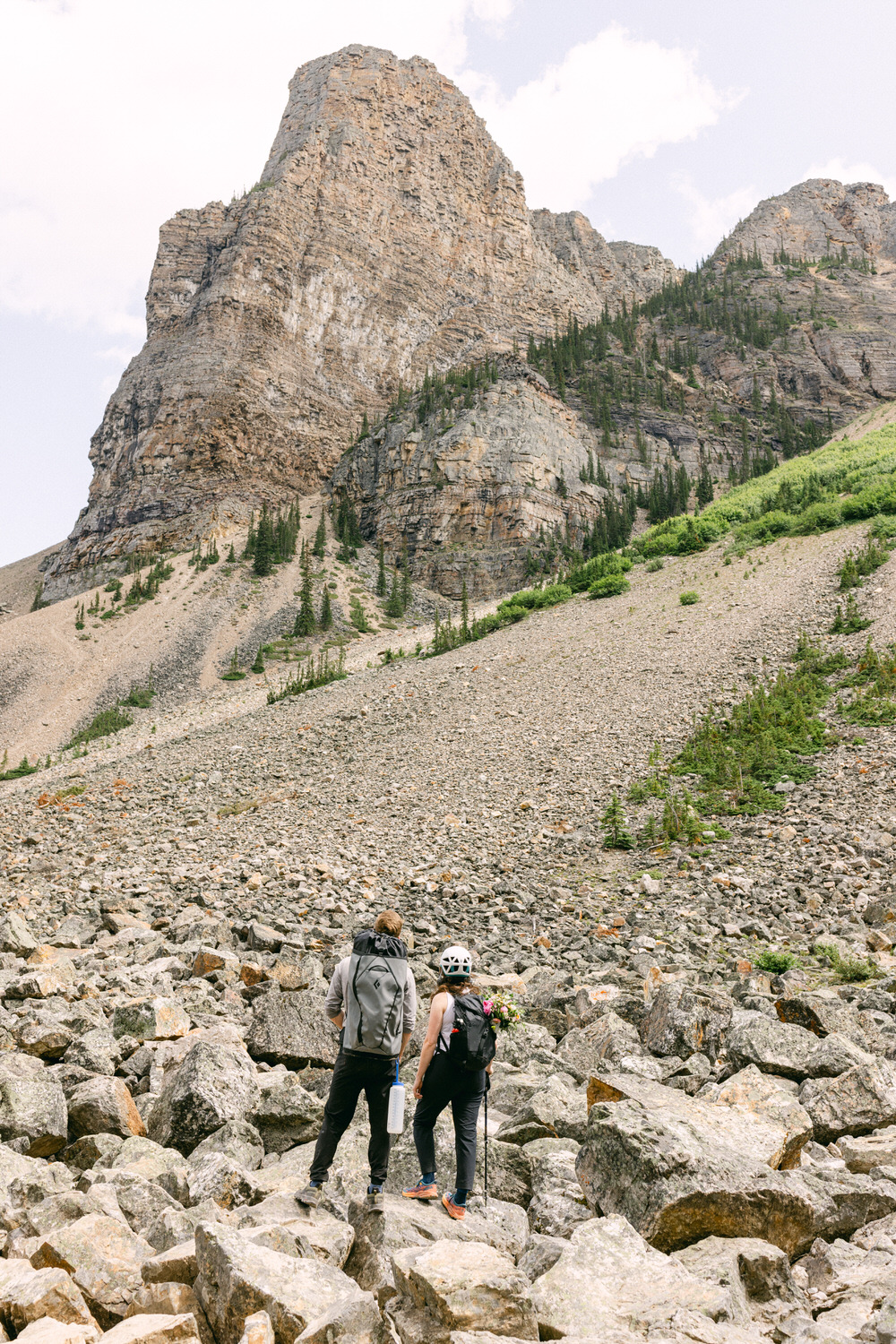 Two hikers navigate through a rocky landscape with towering mountains and greenery in the background.