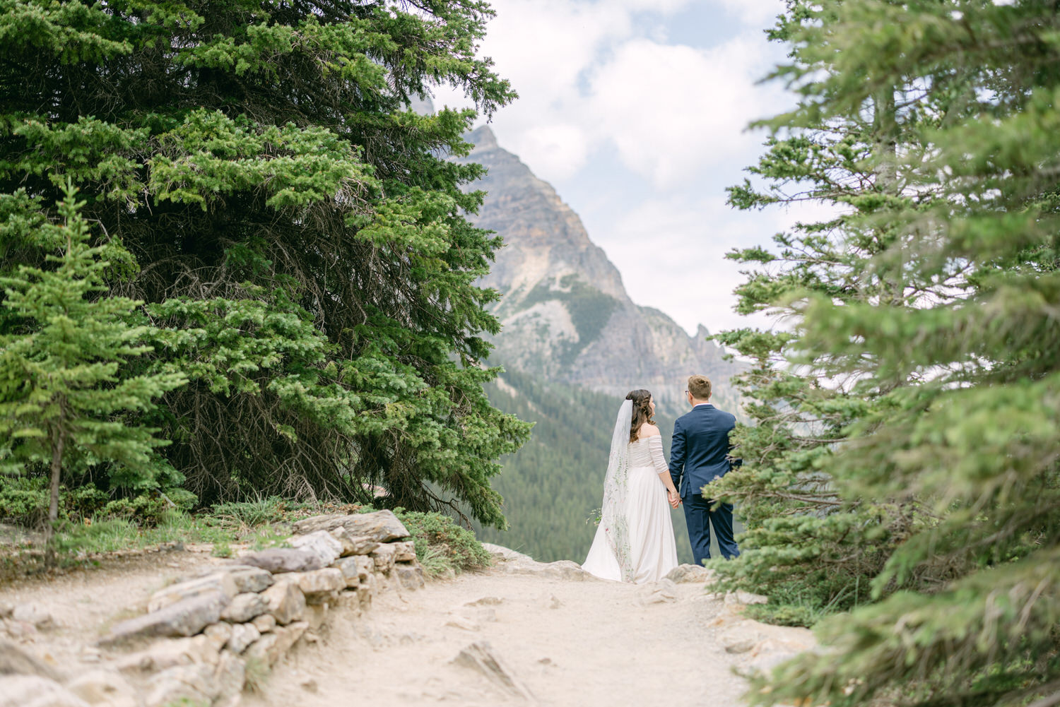 A couple in wedding attire holding hands, standing on a scenic path surrounded by trees and mountains.