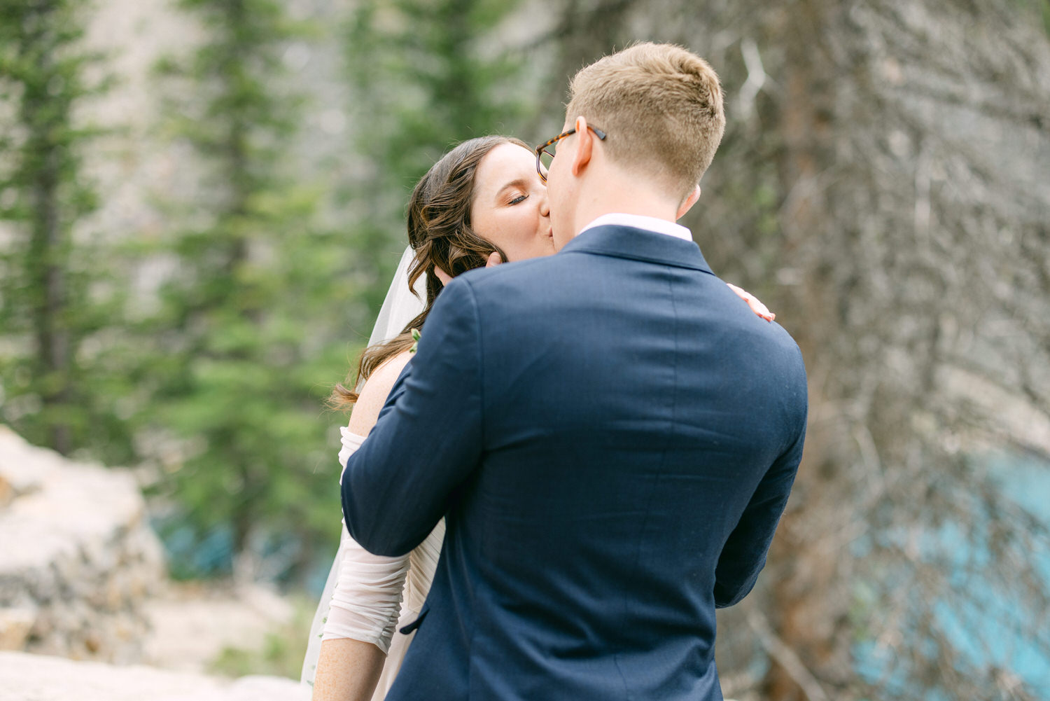 A couple sharing a kiss in a forest setting, with the bride in a strapless dress and veil, and the groom in a navy suit.