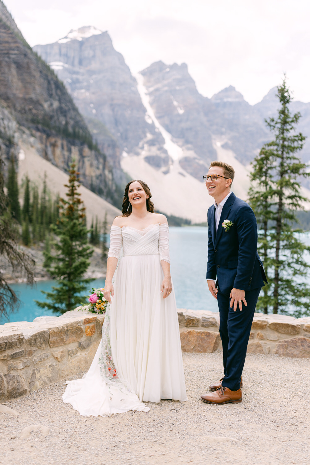 A joyful couple laughing together with a scenic mountain backdrop and a lake, surrounded by trees, during their wedding ceremony.