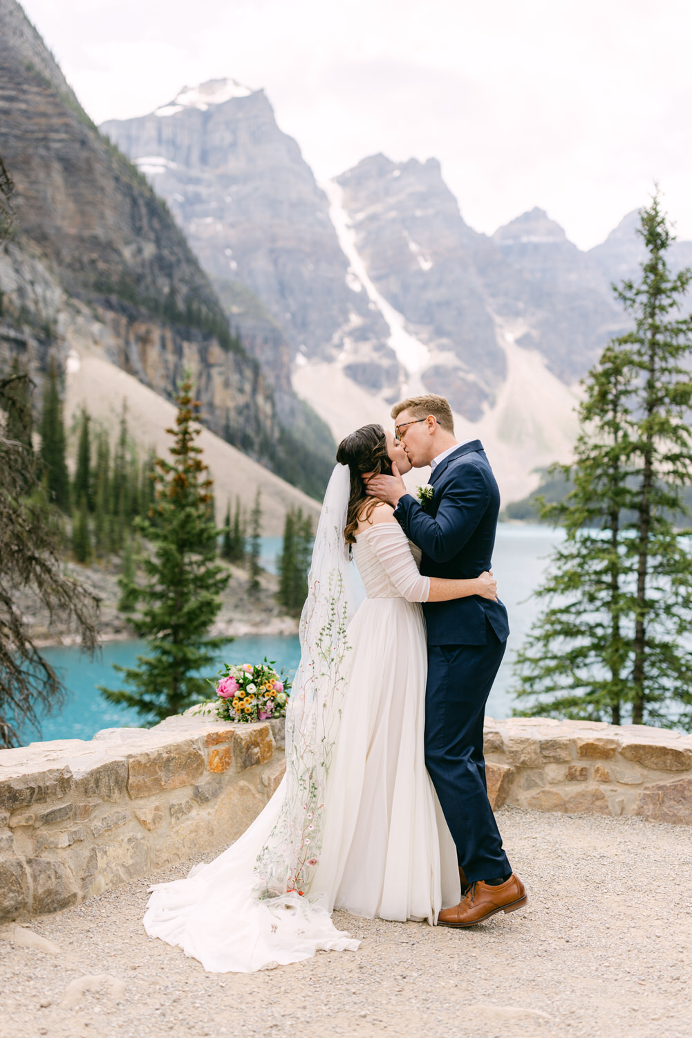 A couple shares a kiss in a picturesque outdoor setting with mountains and a turquoise lake in the background, showcasing love and nature.