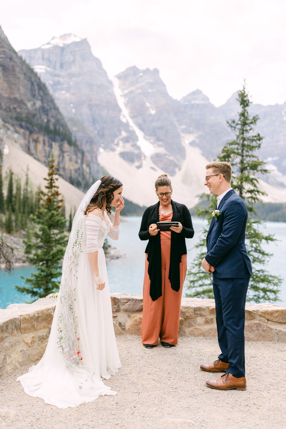 A bride and groom exchange vows in front of a stunning mountain backdrop, with a celebrant officiating, while the bride appears emotional.