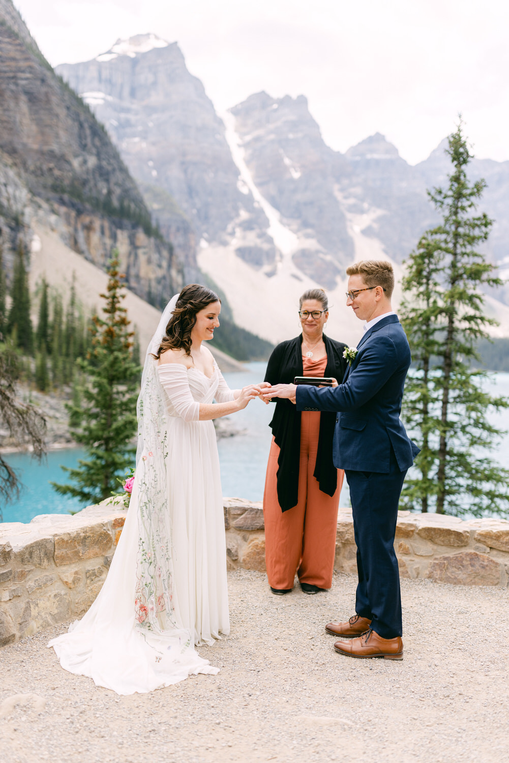 A couple exchanging rings during an outdoor wedding ceremony by a lake, surrounded by mountains and trees.