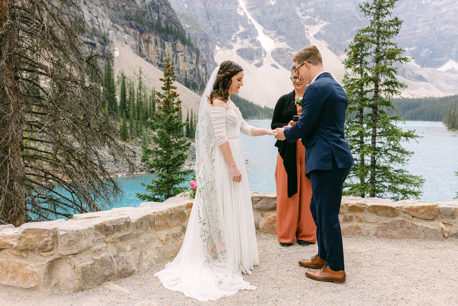 A bride and groom exchange vows by a serene lake surrounded by towering trees and mountains, with an officiant leading the ceremony.