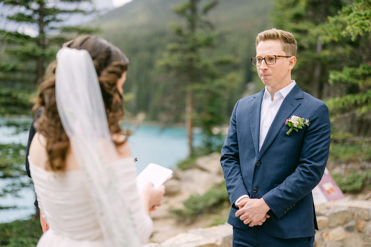 A bride reads her vows to a groom during an outdoor wedding ceremony by a serene lake, surrounded by trees and mountains.